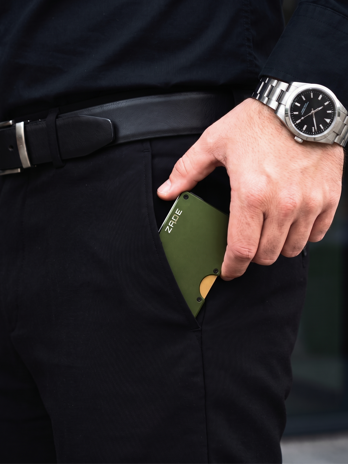 Zade olive green wallet placed into black trouser pocket, hand with steel watch
